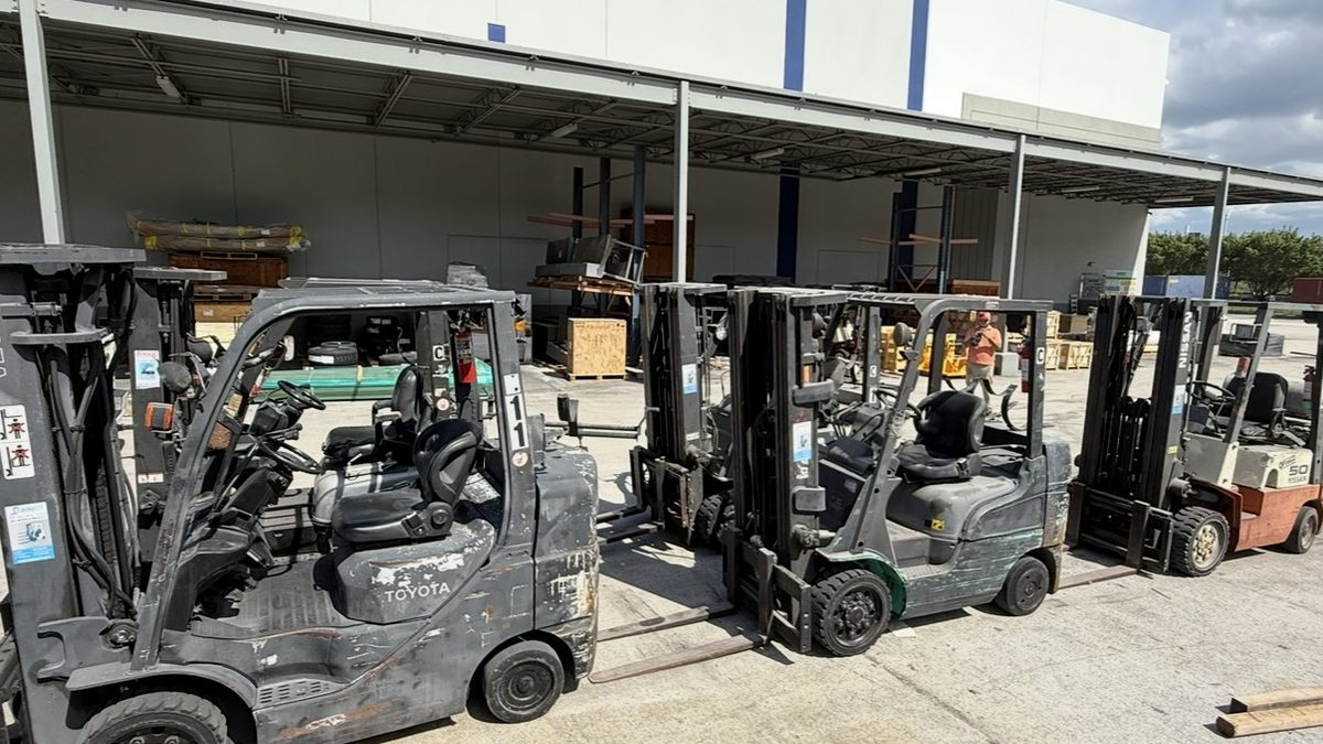 Row of used warehouse forklifts parked outside a distribution building during a fleet buyout