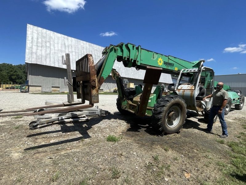 Green telehandler forklift with loose forks and attachments laid out on the ground before removal